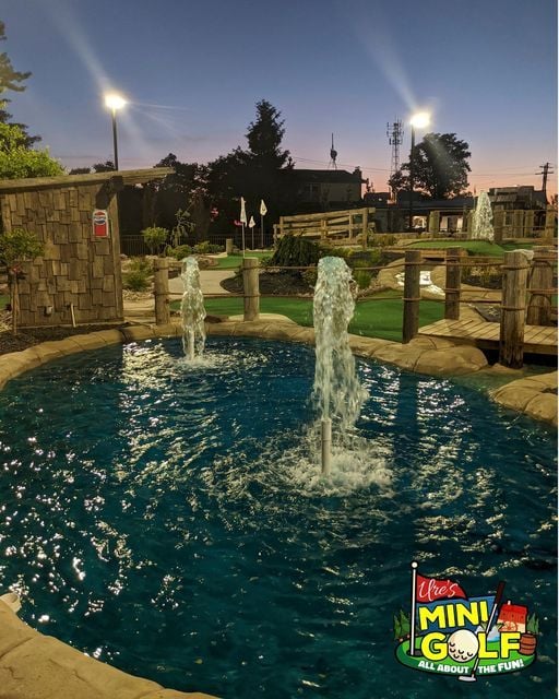 An early evening view onto Ure's Mini-Golf, looking across the pond that features fountains of water shooting up into the sky and the lights for the golf course shimmering in the background