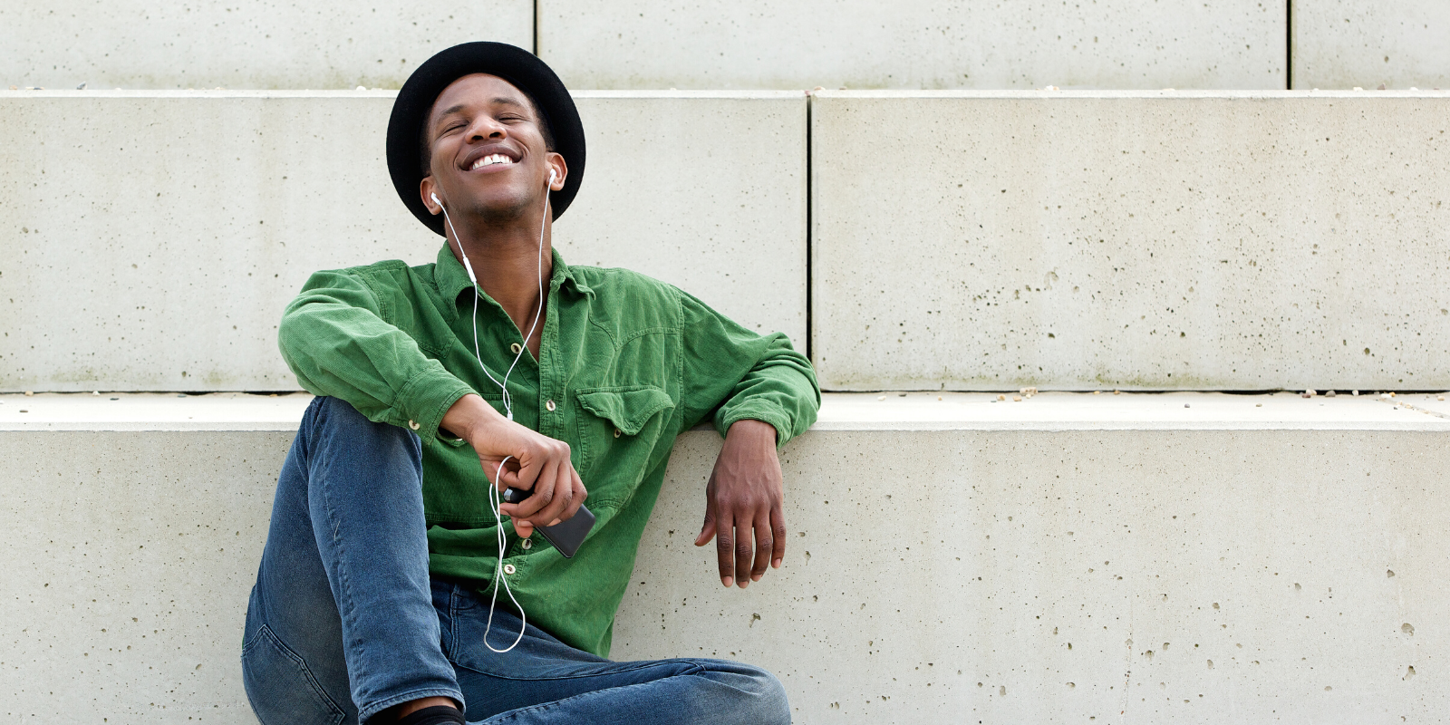 Man in his 30s looking relaxed and happy sitting on the ground in an urban setting with earbuds in