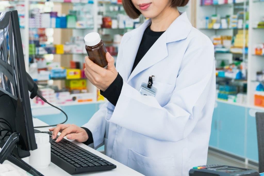 A female compounding pharmacist is holding a bottle of medicine in front of a computer
