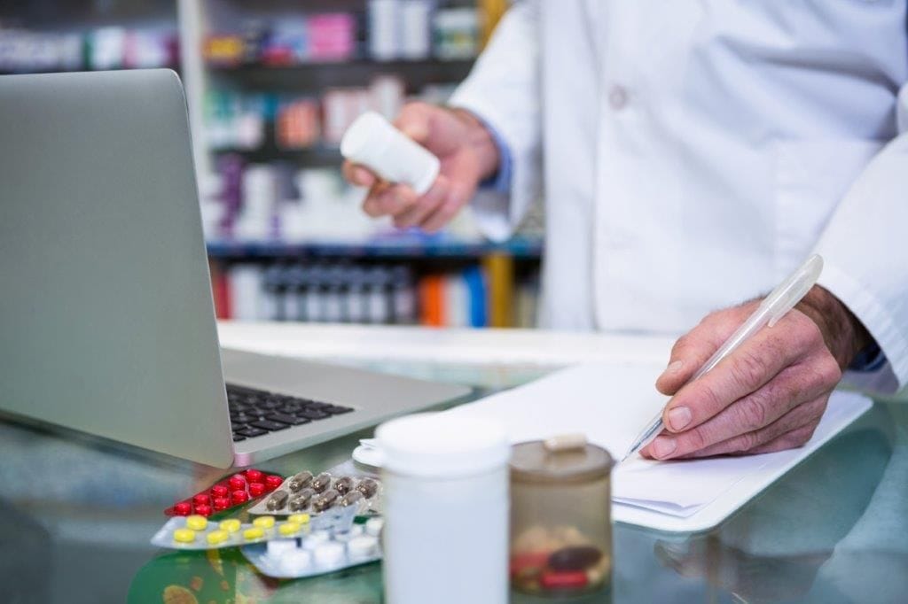 A compounding pharmacist writing ingredients note beside a laptop inside compounding pharmacy.